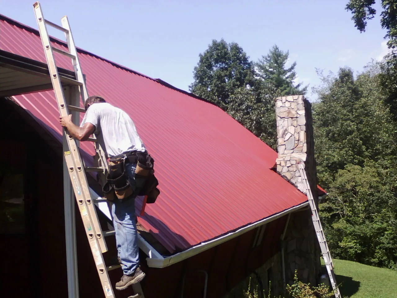 Roofer on a ladder beside a red metal roof and stone chimney — Soffit & Fascia Repair by Roofing Brothers in Millen